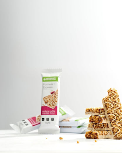 Fire uinnpakkede Herbalife Formula 1 Expressbarer med tranebær og hvit sjokolade er stablet på et bord og en bar lenes mot stabelen. I den litt uklare bakgrunnen ses flere innpakkede Expressbarer med samme smak.