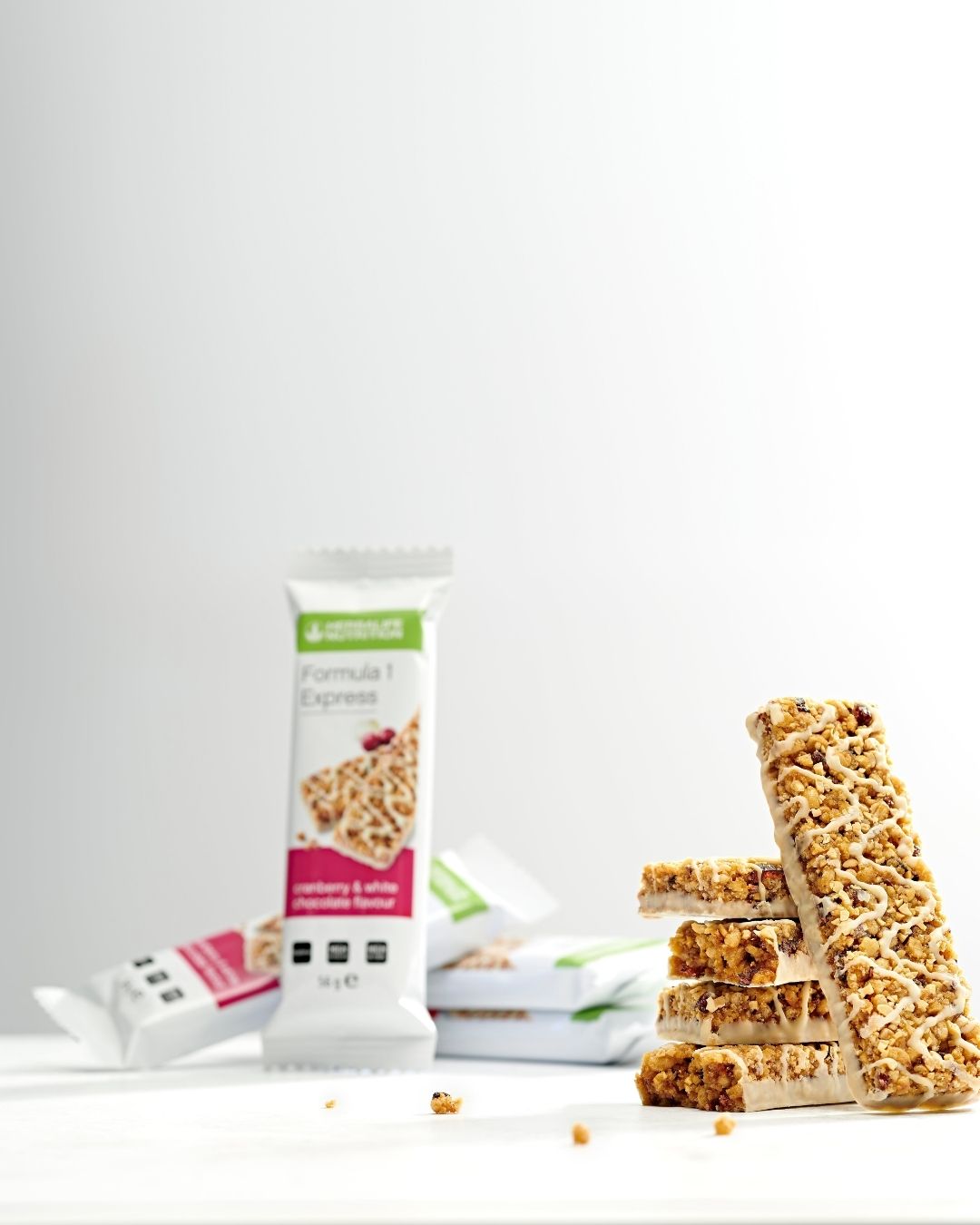Fire uinnpakkede Herbalife Formula 1 Expressbarer med tranebær og hvit sjokolade er stablet på et bord og en bar lenes mot stabelen. I den litt uklare bakgrunnen ses flere innpakkede Expressbarer med samme smak.