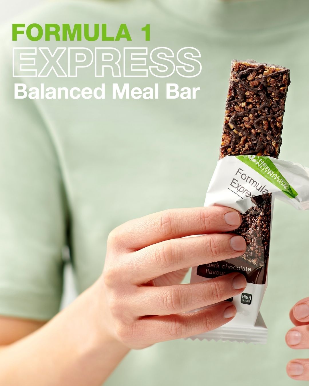 En person i lys grønn t-skjorte holder en åpnet Herbalife Formula 1 mellombar med mørk sjokolade. På bildet står teksten "Formula 1 Express Balanced Meal Bar".