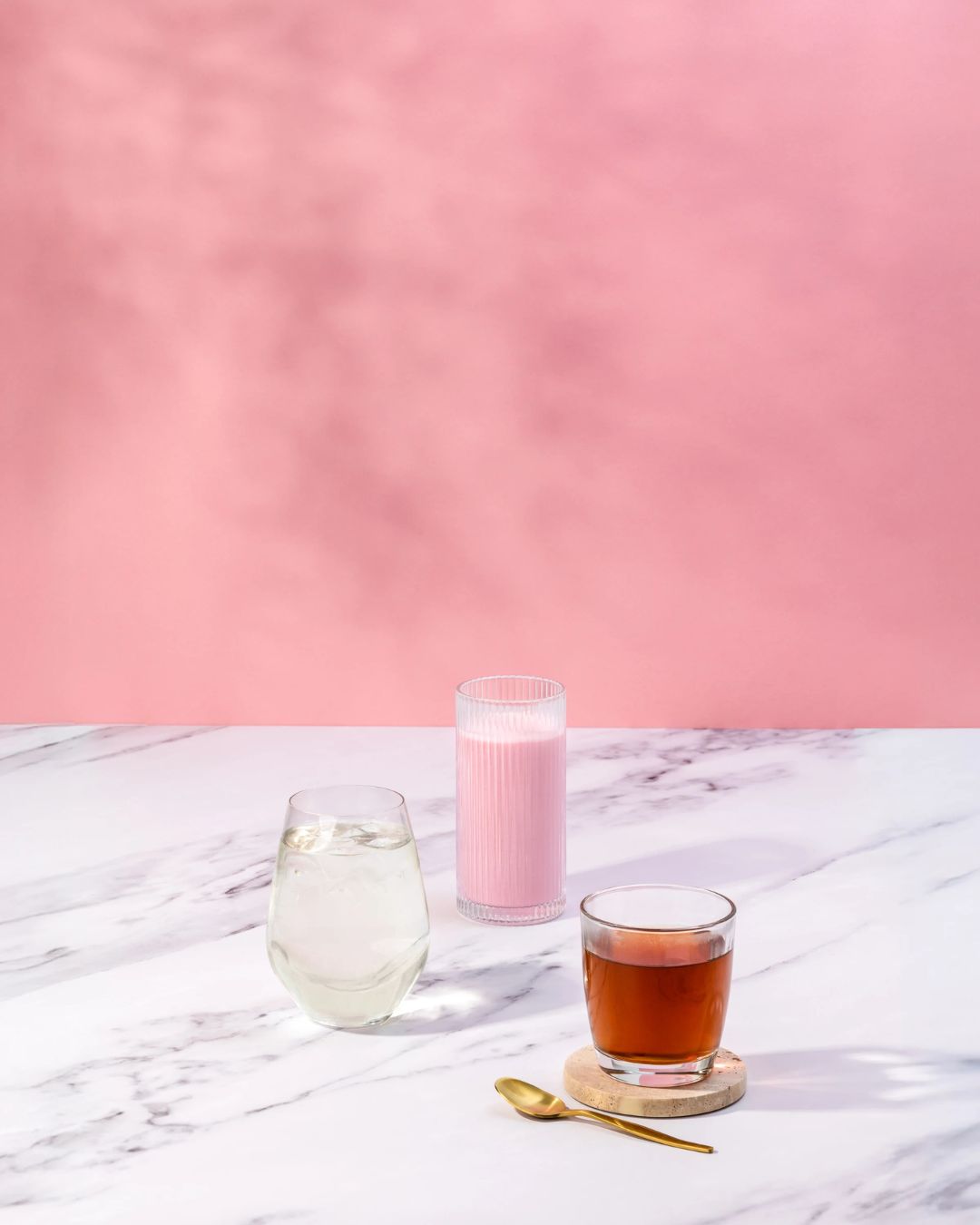 En rosa Herbalife-smoothie i et glass står på et marmorbord sammen med et glass aloe vera-juice og en kopp te. Rosa vegg bak.