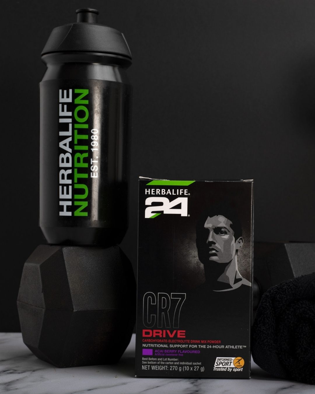 En pakke Herbalife H24 CR7 Drive porsjonspose og en løs porsjonspose står sammen med en sportsdrikkeflaske med Herbalife-logo og noen dumbbells. 