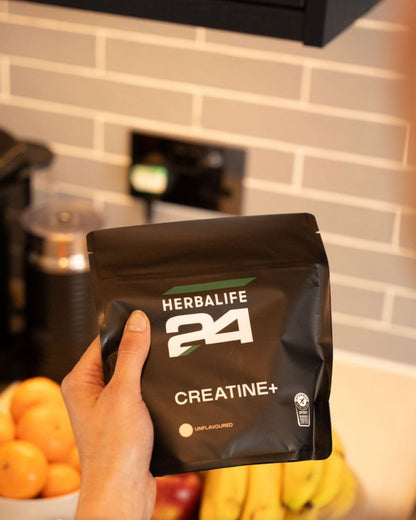 En hånd holder en pose Herbalife H24 Creatine+ kreatin monohydrat, i bakgrunnen ses ulike frukter på en kjøkkenbenk