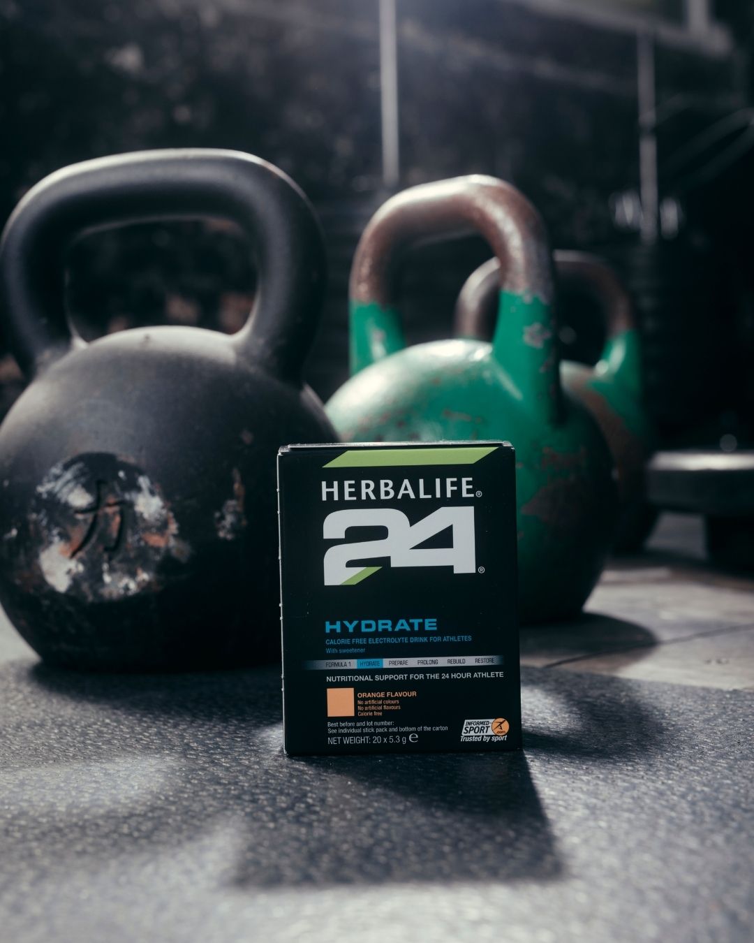 En pakke Herbalife H24 Hydrate isotonisk sportsdrikk står på gulvet foran noen kettlebells. 