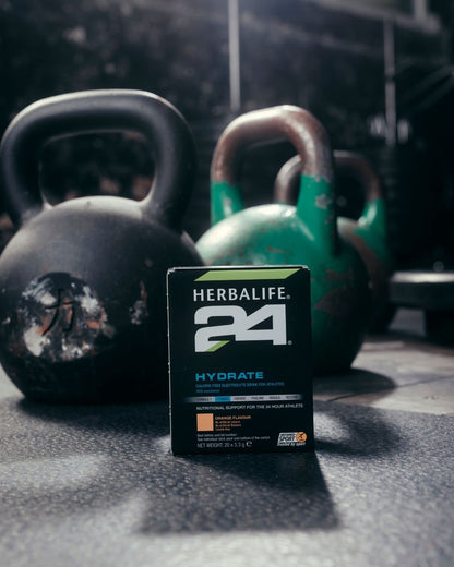 En pakke Herbalife H24 Hydrate isotonisk sportsdrikk står på gulvet foran noen kettlebells. 