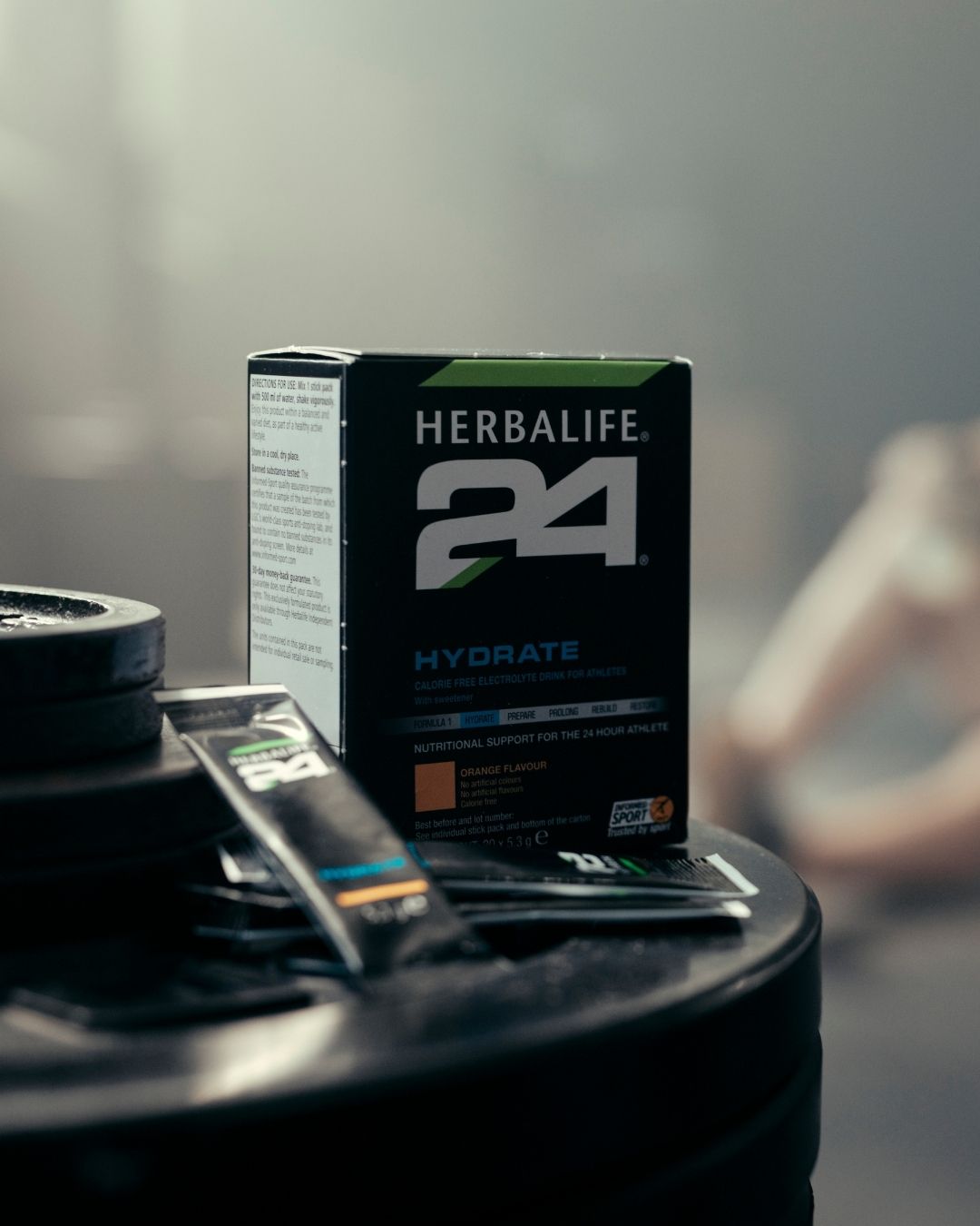 En pakke Herbalife H24 Hydrate isotonisk sportsdrikk og to løse porsjonsposer står på en stabel vektskiver. Utydelig i bakgrunnen ses en mann i treningstøy. 