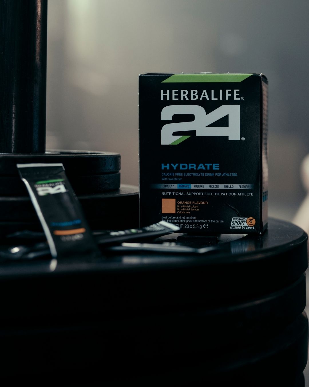 En pakke Herbalife H24 Hydrate isotonisk sportsdrikk og to løse porsjonsposer står på en stabel vektskiver. 