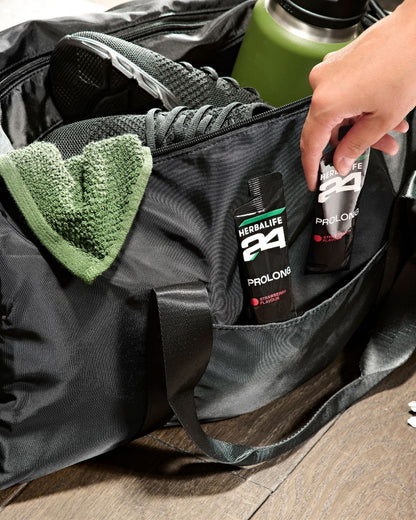 En åpen treningsbag med sko og drikkeflaske og en hånd som tar en av to poser Herbalife H24 Prolong Energy Gel fra den.