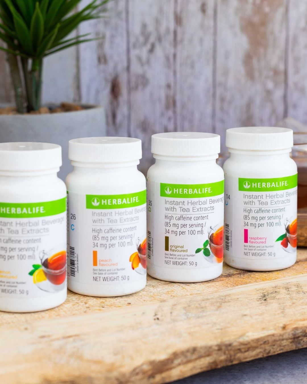 Fire bokser Herbalife Instant urtekonsentrat te med de ulike smakene sitron, fersken, original og bringebær på en treplanke. I bakgrunnen skimtes en kopp urtete. 