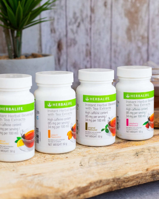 Fire bokser Herbalife Instant urtekonsentrat te med de ulike smakene sitron, fersken, original og bringebær på en treplanke. I bakgrunnen skimtes en kopp urtete. 