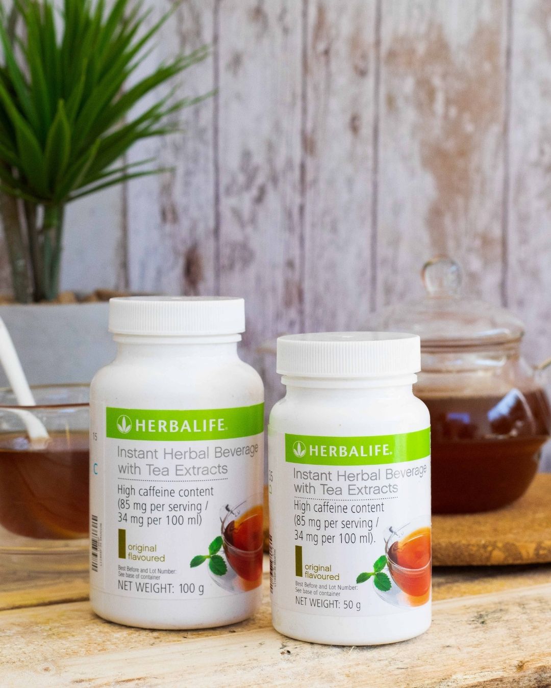 En stor og en liten boks Herbalife Instant urtekonsentrat te med originalsmak. Bak står en glasskopp med te og ei tekanne i glass med te. 