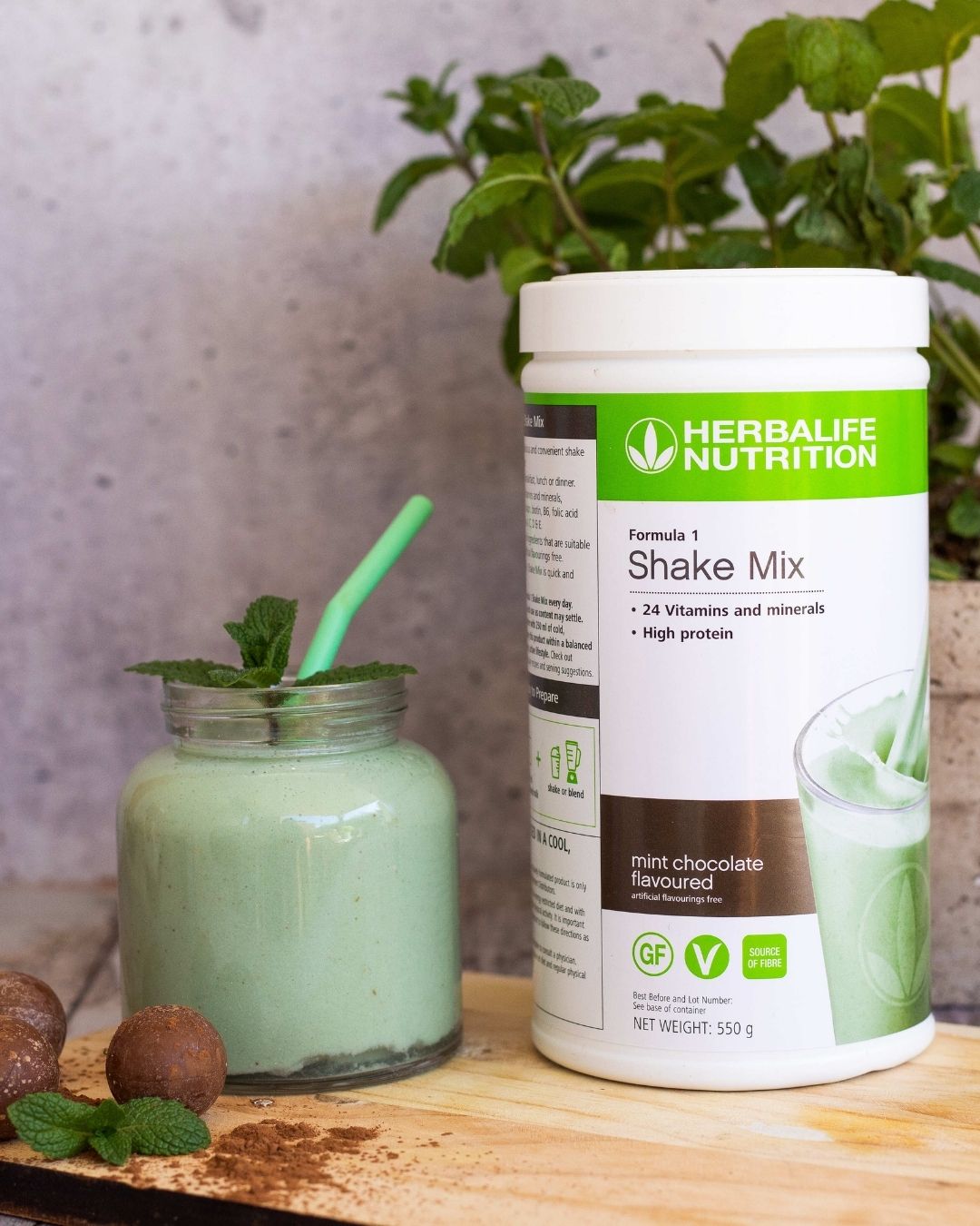 En boks Herbalife Formula 1 shakemix med Mint chocolate-smak sammen med et glasskrus med mintsjokolade-shake og et mintgrønt sugerør. Tre sjokoladekuler strødd med kakao og et mintblad ligger foran glasset. I bakgrunnen ses en mynteplante.
