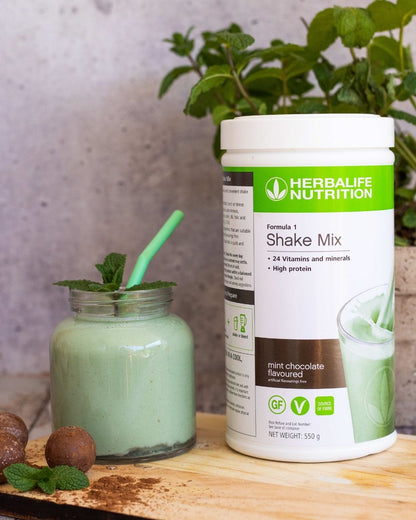 En boks Herbalife Formula 1 shakemix med Mint chocolate-smak sammen med et glasskrus med mintsjokolade-shake og et mintgrønt sugerør. Tre sjokoladekuler strødd med kakao og et mintblad ligger foran glasset. I bakgrunnen ses en mynteplante.