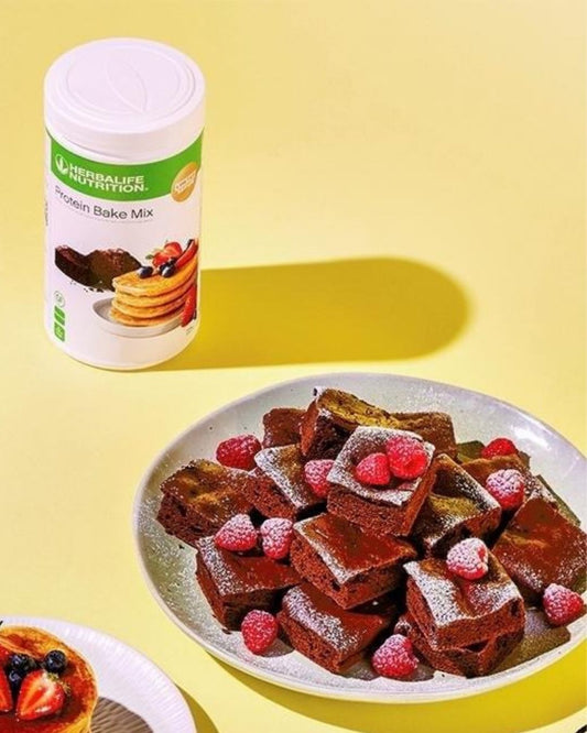En boks Herbalife Protein Bake Mix sammen med eksempler på ulike retter laget med bakemiksen; brownies og pannekaker.