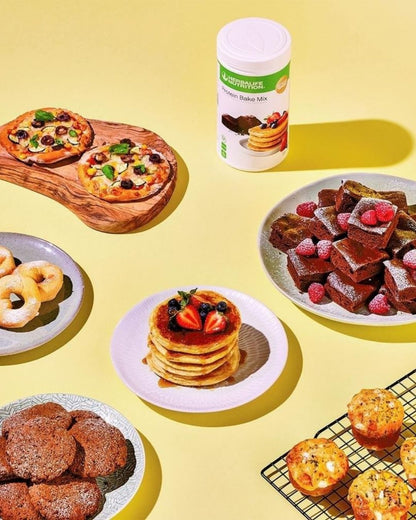 En boks Herbalife Protein Bake Mix sammen med eksempler på ulike retter laget med bakemiksen; pizza, smultringer, sjokoladecookies, amerikanske pannekaker, brownies og muffins.