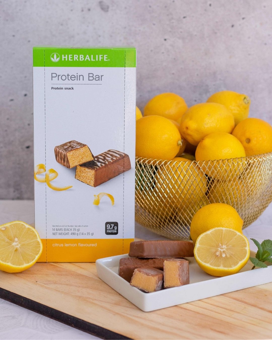 Proteinbarer med sitronsmak fra Herbalife i eske står på ei trefjøl sammen med et fat med åpnede proteinbarer og noen sitroner.