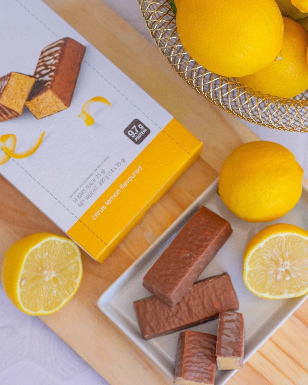 Proteinbar med sitronsmak fra Herbalife i eske og 3 åpnede barer uten papir ligger på ei lys trefjøl sammen med noen sitroner og en myntekvast.