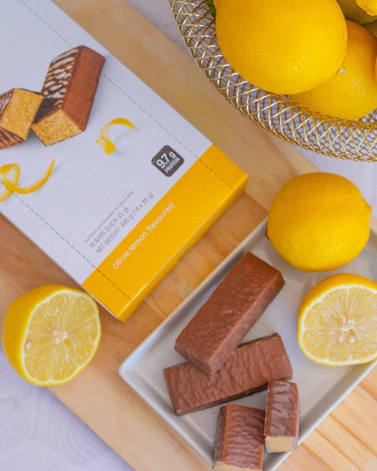 Proteinbar med sitronsmak fra Herbalife i eske og 3 åpnede barer uten papir ligger på ei lys trefjøl sammen med noen sitroner og en myntekvast.