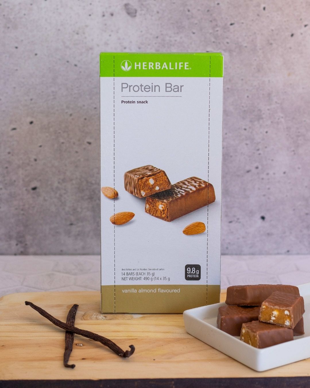 En eske Herbalife proteinbarer med vanilje-mandel-smak på ei trefjøl sammen med et fat åpnede proteinbarer og to vaniljestenger.