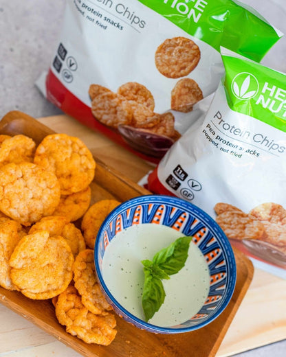 To porsjonsposer Herbalife Protein Chips proteinrik snacks med barbeque-smak, står på en lys trefjøl sammen med et fat med proteinchips og en skål med dip.