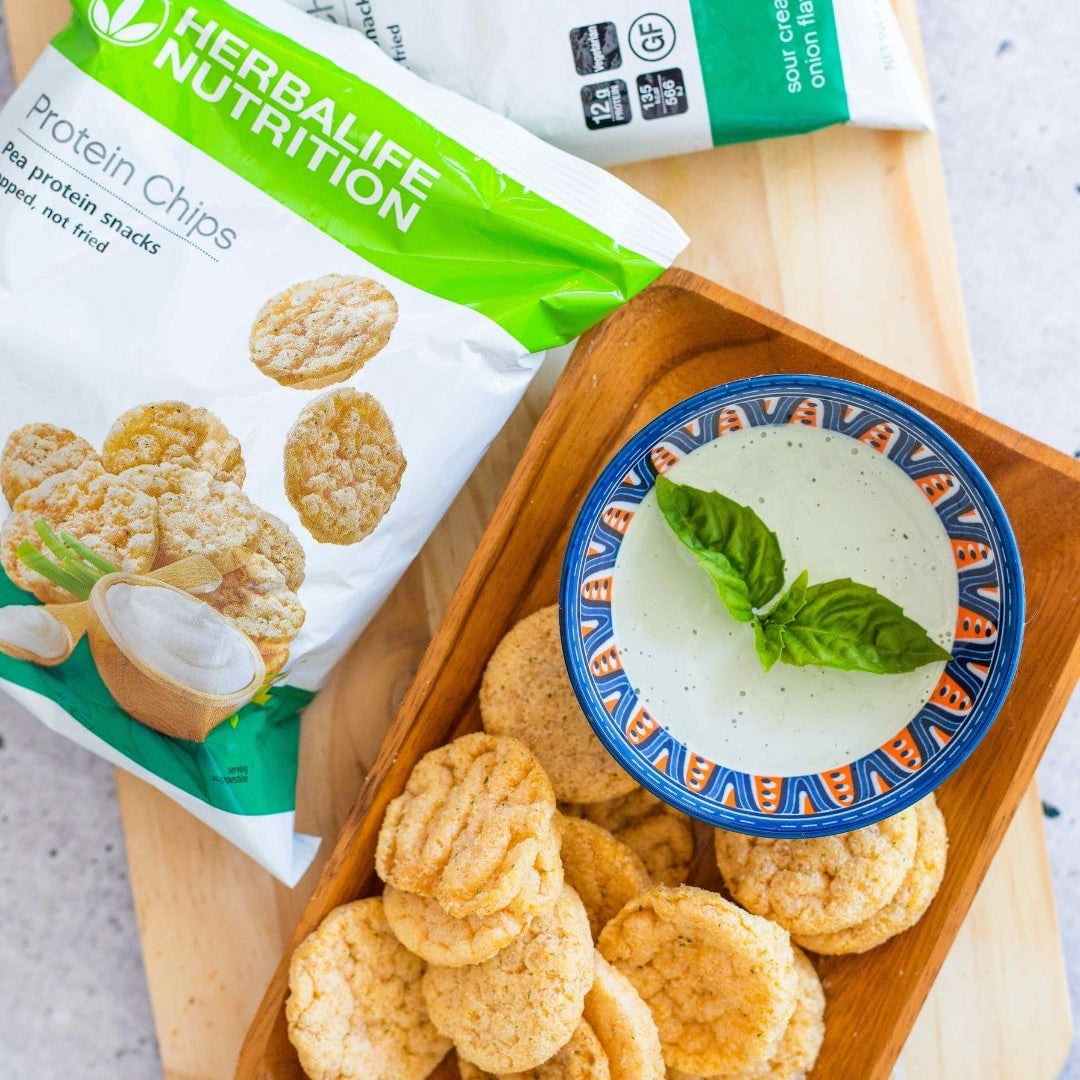 En porsjonspose Herbalife Protein Chips proteinrik snacks sourcream & onion-smak ligger på ei lys trefjøl sammen med et fat med proteinchips og ei skål med dip.