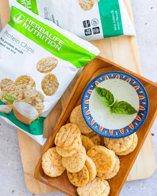 En porsjonspose Herbalife Protein Chips proteinrik snacks sourcream & onion-smak ligger på ei lys trefjøl sammen med et fat med proteinchips og ei skål med dip.