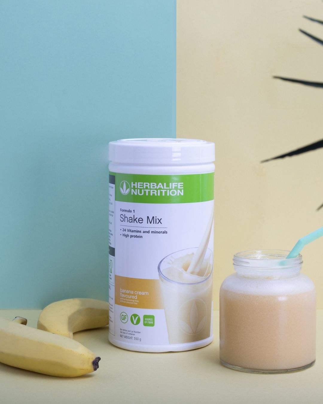 En boks Herbalife banan-shake, et glasskrus med banan-smoothie og mintgrønt sugerør. To uskrelte gule bananer og en grønn plante i bakgrunnen.