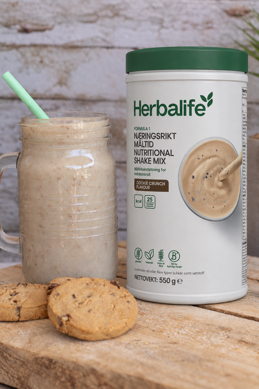 Herbalife Formula 1 shakemix med Cookie Crunch-smak og en smoothie på ei lys trefjøl. Smoothien serveres med sugerør i et glasskrus. I forgrunnen tre cookies med sjokoladebiter. 