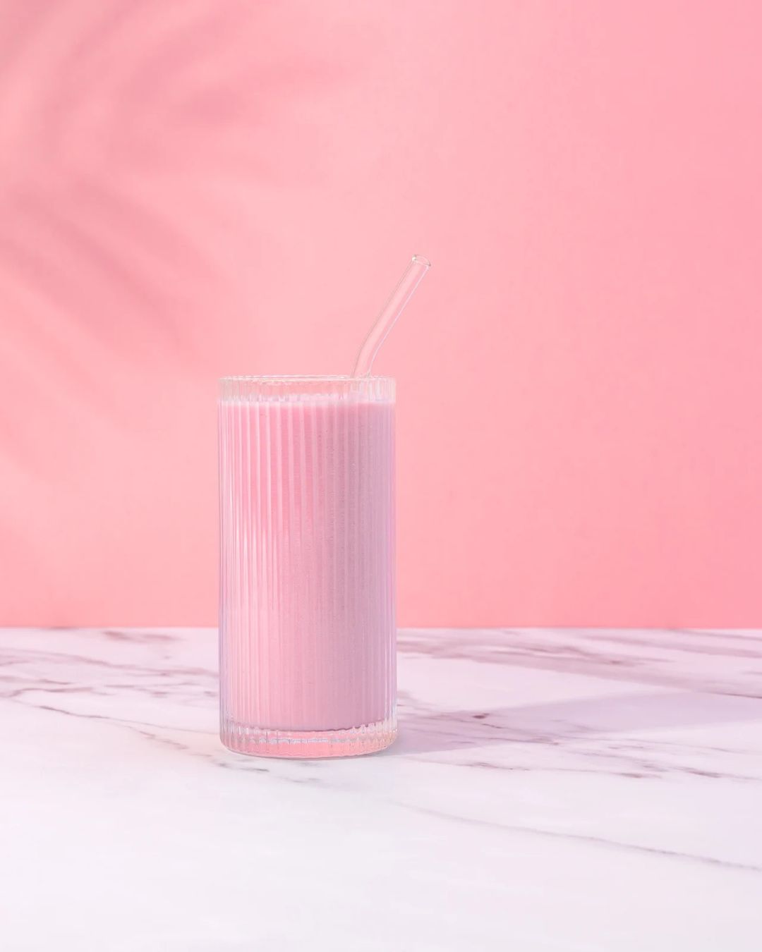 En rosa Herbalife-smoothie i et glass står på et marmorbord. Rosa vegg bak og det ses en skygge av et palmeblad på veggen.