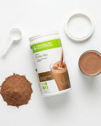 Herbalife shakeboks med sjokoladesmak ligger på et bord med pulver, måleskje og et glass smoothie. 