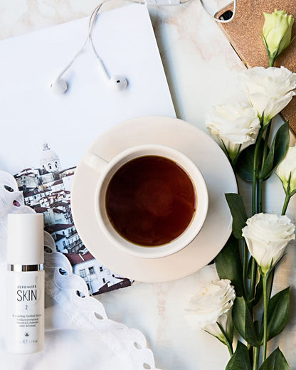 En sprayflaske Herbalife SKIN Energising Herbal Toner ansiktsvann på et bord sammen med en kopp te, en mobiltelefon med headset og noen hvite blomster.  