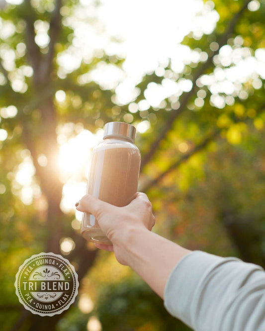 En kvinne holder en Herbalife TriBlend-shake opp i lufta ute i naturen. 