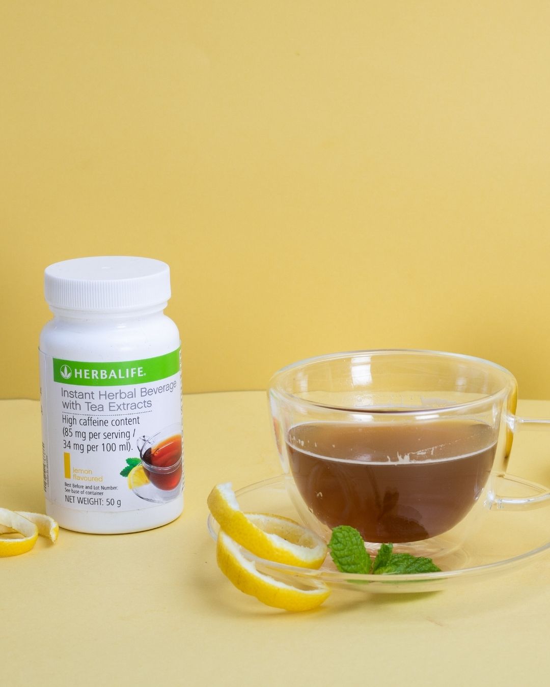 En glasskopp sitronte med sitronskall og mynteblad på skåla. En boks Herbalife Instant Lemon urtekonsentrat te med sitronsmak, 51 gram.