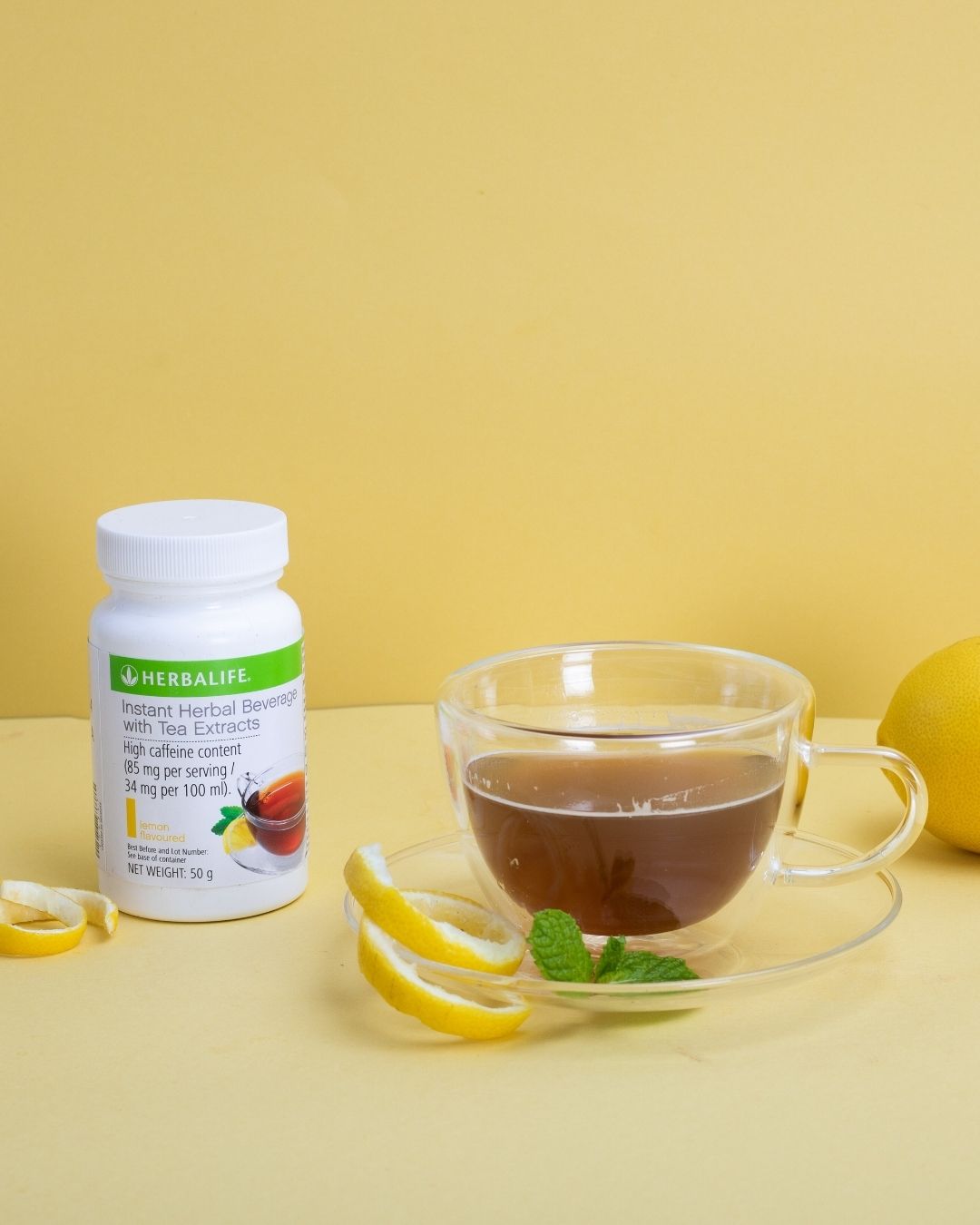 En glasskopp sitronte med sitronskall og mynteblad på skåla. En hel sitron. En boks Herbalife Instant Lemon urtekonsentrat te med sitronsmak, 51 gram.