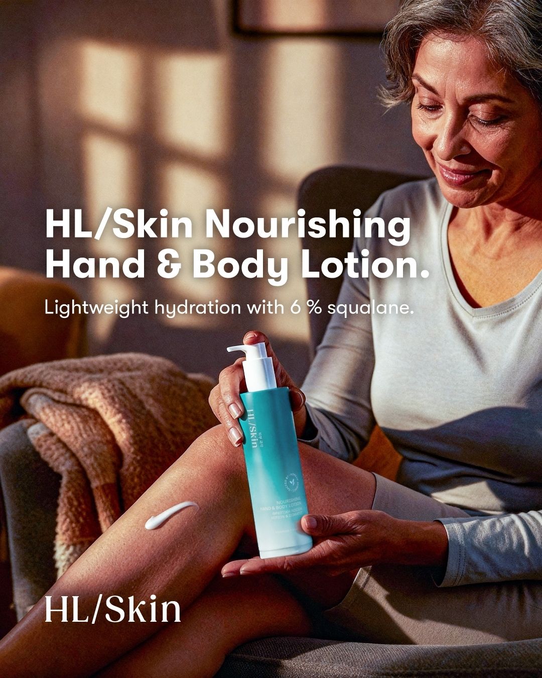 En dame med grått hår og grå t-skjorte har body lotion på leggen klar til å smørs utover. Tekst på bildet: HL/Skin Nourishing Hand & Body Lotion. Lightweight hydration with 6% squalane. 