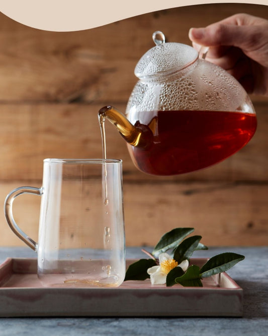 Rykende varm Herbalife Instant Urtete helles fra en glasskanne i en gjennomsiktig kopp, på et fat med dekorativer blader og blomst ved siden av. Perfekt for en sunn og oppkvikkende start på dagen. 