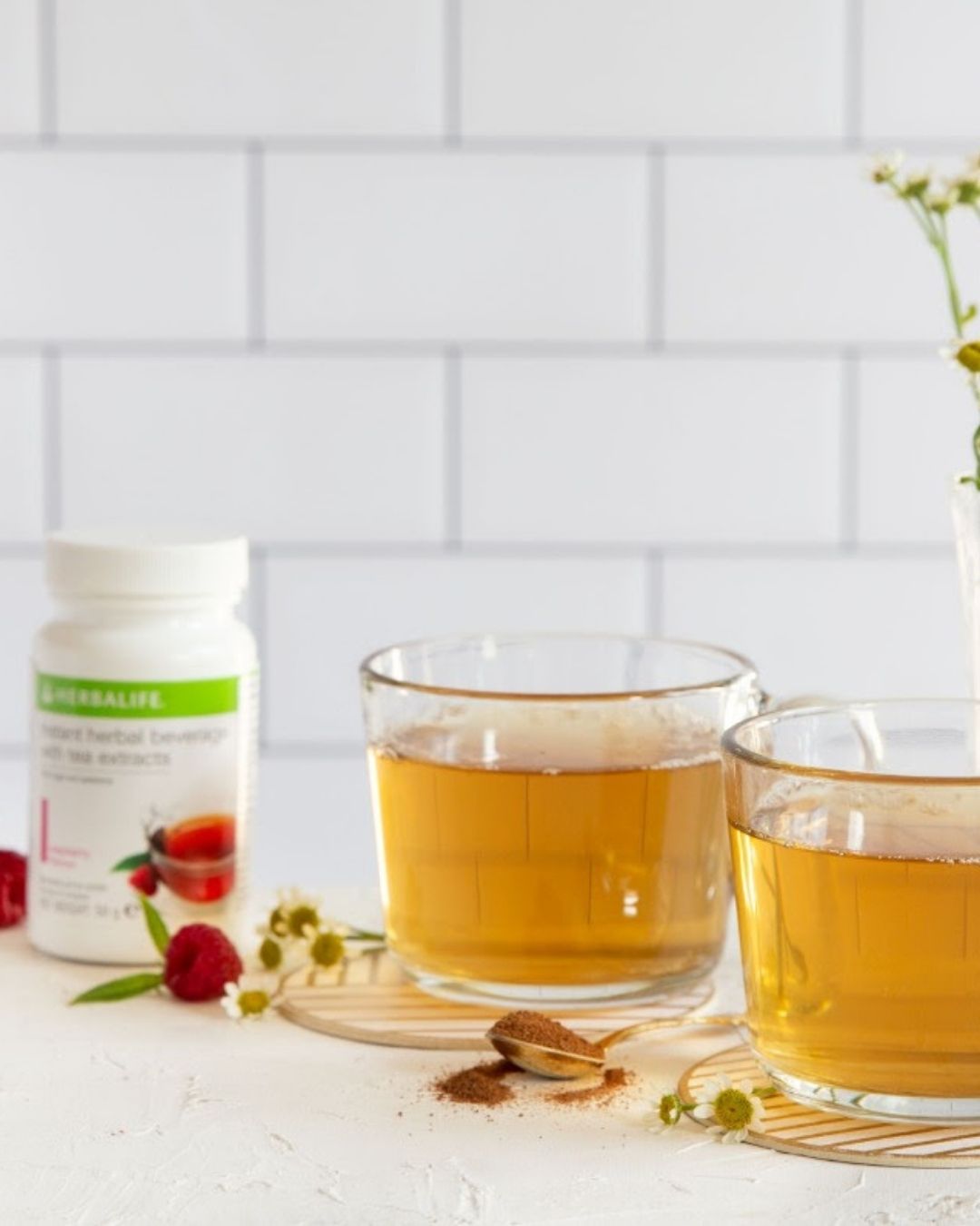 To glasskopper bringebær-te foran en vase med hestehov, et ferskt bringebær på bordet ligger sammen med en teskje instant te. I bakgrunnen ses en boks Herbalife urtekonsentrat raspberry. 