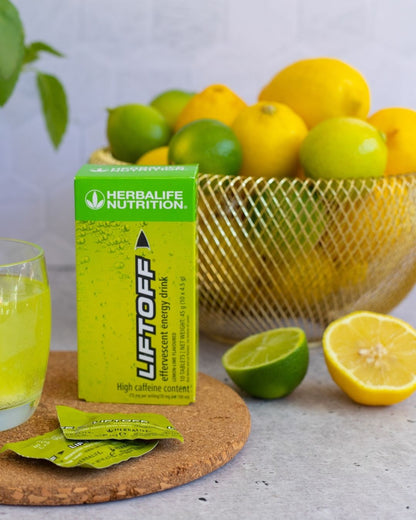 En pakke Herbalife Liftoff energidrikk står sammen med et glass liftoff, to løse porsjonspakker og en skål med lime og sitroner. 