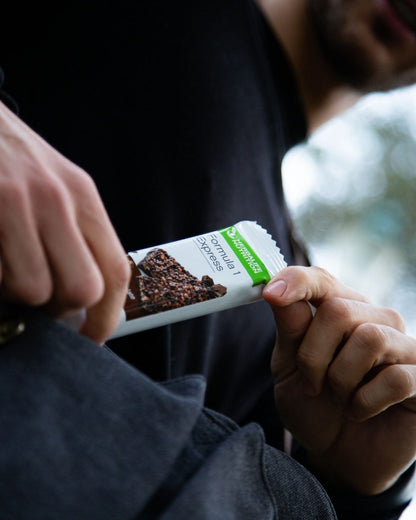 En person i en mørkeblå jakke tar en Herbalife Formula 1 mellombar med mørk sjokolade opp av jakkelomma.