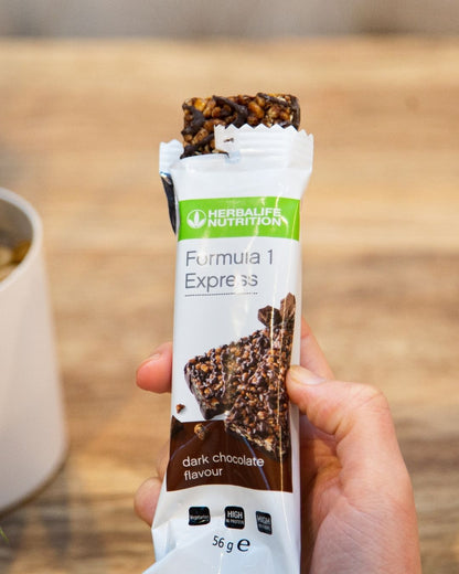 En Herbalife Formula 1 Expressbar halveis trukket ut av papiret sitt og en hånd som holder den. 