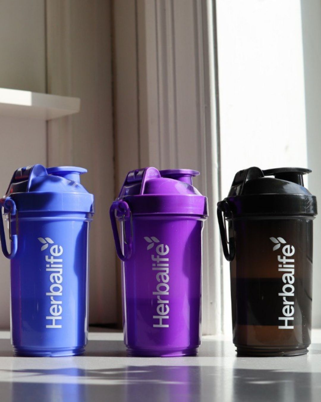Tre Herbalife Smartshake-flasker i blå, lilla og svart, hver med hvit Herbalife-logo og karabinkrok, plassert på et bord ved et vindu.