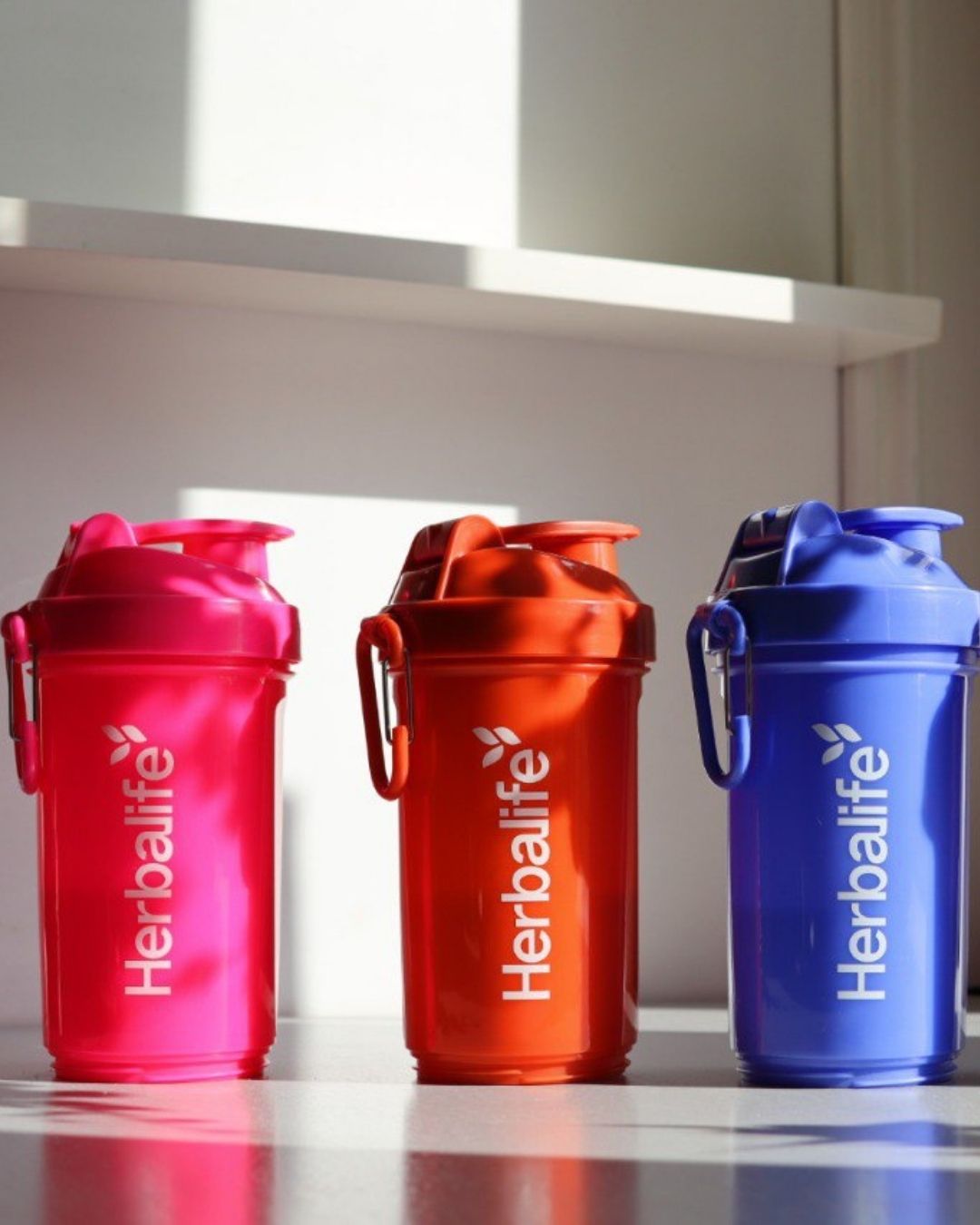 Tre Herbalife Smartshake-flasker på rekke i rosa, oransje og blå farge, hver med hvit Herbalife-logo og karabinkrok.